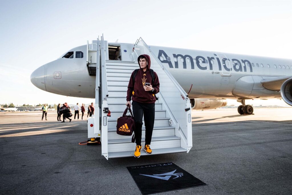 An ASU student athlete in an ASU-branded maroon hoodie and brown pants descends an American Airlines airplane stairway, holding a smartphone in one hand and a maroon Adidas duffel bag in the other.