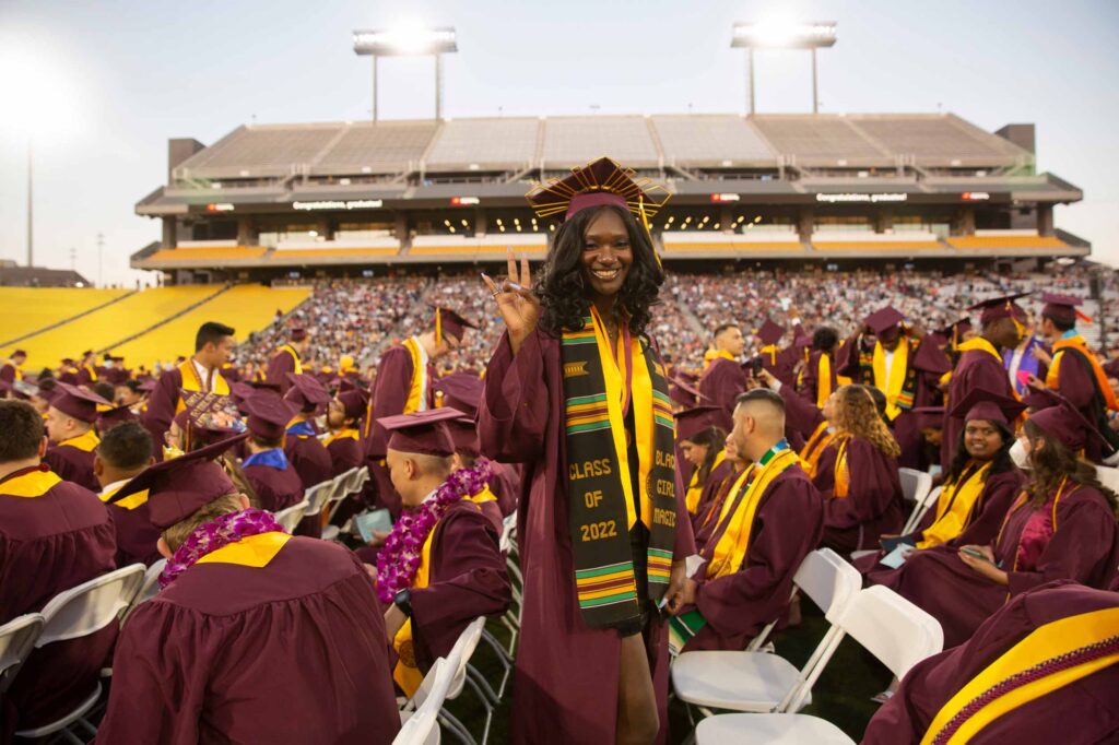 A African-American woman graduate shows the ASU pitchfork gesture while she poses for a photo in her full regalia at a Fulton Schools Convocation ceremony in ASU's Sun Devil Stadium.